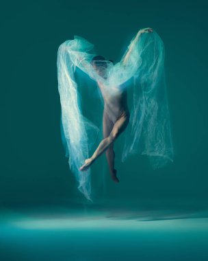 Jump and hover while in air. Flexible ballet dancer in motion with white cloth, fabric isolated on green blue background. Art, beauty, creation, inspiration concept. Blurring effect