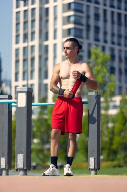 Rest time after active strength training. Handsome young muscular man in red shorts at sports ground at public park in summer morning. Wellness, sport, fitness, healthy lifestyle concept.