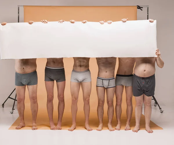 Slender legs. Group of body positive men standing together in underwear. Models holding big blank paper banner closing faces. Body positive concept