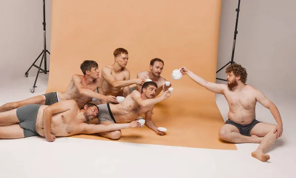 Leadership. Group of happy cheerful men of different ages and body shapes sitting on floor and drinking tea at studio photo shoot. Feeling comfortable. Fashion models shirtless