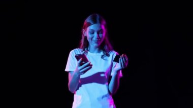Smiling young girl pays for purchase with credit card and mobile app. Dark neoned background. Concept of business, black friday sales, ad, discount, cyber monday and online purchases.