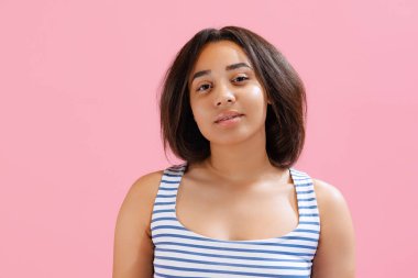 Look calm, composed. Beautiful african girl wearing summer striped tank top and shorts looking at camera isolated on pink background. Beauty, art, fashion, emotions concept