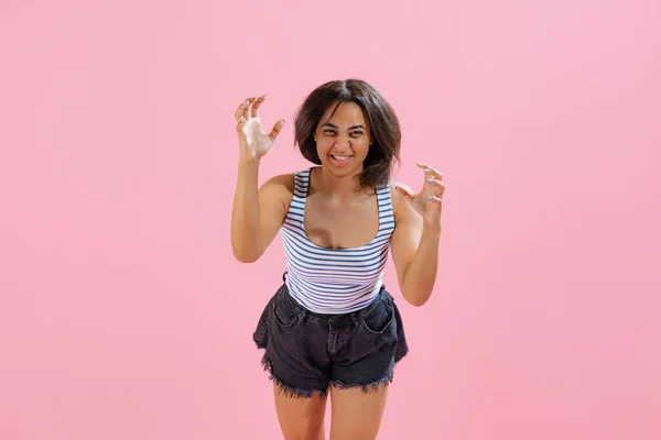 Negative emotions. Angry. Beautiful african girl wearing summer striped tank top and shorts looking at camera isolated on pink background. Beauty, art, fashion, emotions concept