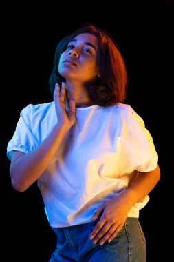 Thoughts. Young charming african girl, student in white t-shirt posing isolated on dark background in neon light. Concept of beauty, art, fashion, education, youth and ad