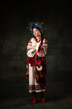 Sadness. Art portrait of beautiful woman wearing traditional folk Ukrainian costume posing isolated over dark vintage background. Fashion, beauty, cultural heritage concept.