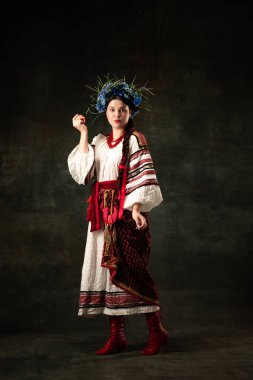 Emotional adorable woman wearing national folk Ukrainian attire posing isolated over dark vintage background. Fashion, beauty, cultural heritage concept.