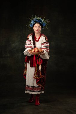 Hospitality. Art portrait of beautiful woman wearing traditional folk Ukrainian costume posing isolated over dark vintage background. Fashion, beauty, cultural heritage concept.