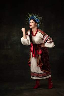 Figth for rights. Emotional adorable woman wearing national folk Ukrainian attire posing isolated over dark vintage background. Fashion, beauty, cultural heritage concept.