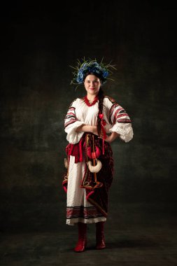 Emotional adorable woman wearing national folk Ukrainian attire posing isolated over dark vintage background. Fashion, beauty, cultural heritage concept.