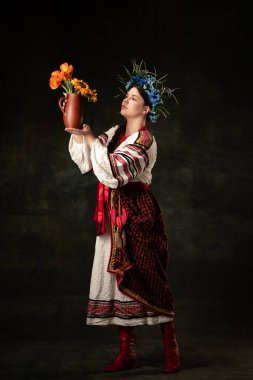 Art portrait of beautiful woman wearing traditional folk Ukrainian costume posing with flowers isolated over dark vintage background. Fashion, beauty, cultural heritage concept.