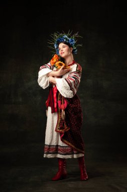 Hope and faith. Art portrait of beautiful woman wearing traditional folk Ukrainian costume posing isolated over dark vintage background. Fashion, beauty, cultural heritage concept.