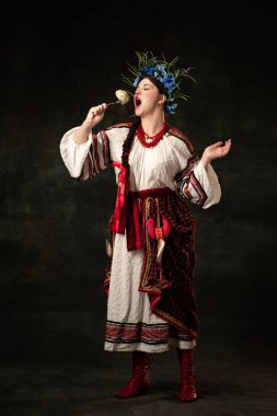 Traditions, national cuisine. Creative portrait of beautiful Ukrainian woman in traditional folk costume tasting dumpling isolated over dark background. Fashion, beauty, cultural heritage concept.