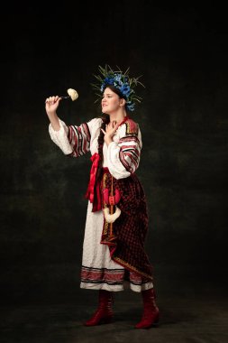 Traditions, national cuisine. Creative portrait of beautiful Ukrainian woman in traditional folk costume tasting dumpling isolated over dark background. Fashion, beauty, cultural heritage concept.