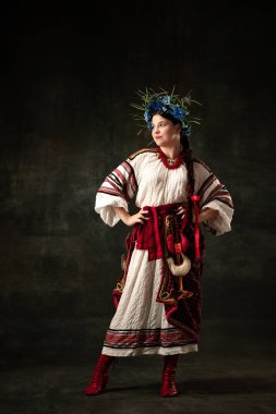 Emotional young woman wearing national folk Ukrainian attire isolated over dark retro background. Human emotions, history, ad, fashion, beauty, eras comparison concept.
