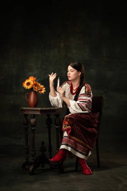 Fortune-telling. Young beautiful Ukrainian woman wearing traditional folk costume isolated over dark vintage background. Fashion, beauty, cultural heritage concept. Human emotions