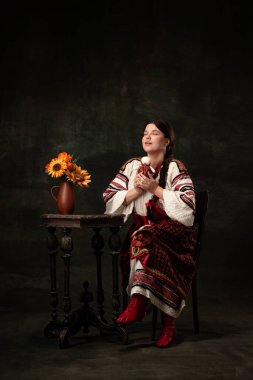 Fortune-telling. Young beautiful Ukrainian woman wearing traditional folk costume isolated over dark vintage background. Fashion, beauty, cultural heritage concept. Human emotions