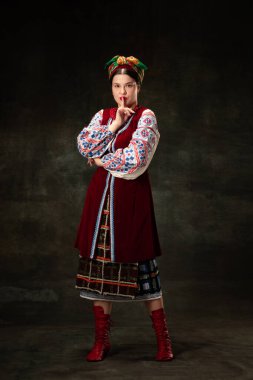 Keep silence. Emotional adorable woman wearing national folk Ukrainian attire posing isolated over dark vintage background. Fashion, beauty, cultural heritage concept.