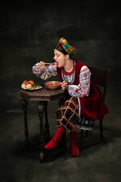 Traditions, national cuisine. Creative portrait of beautiful Ukrainian woman wearing traditional folk costume isolated over dark vintage background. Fashion, beauty, cultural heritage concept.