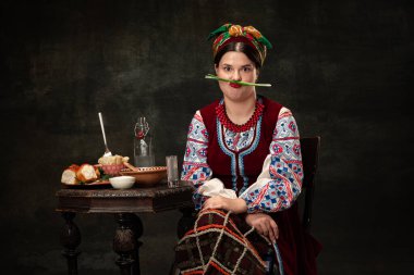 Grimace. Emotional young woman wearing national folk Ukrainian attire isolated over dark retro background. Human emotions, history, ad, fashion, beauty, eras comparison concept.