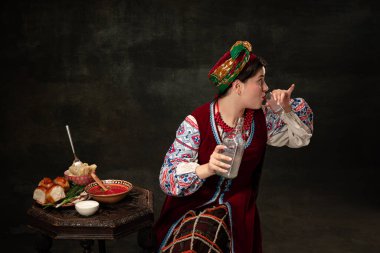 One young beautiful Ukrainian woman in traditional folk Ukrainian costume isolated over dark vintage background. Fashion, beauty, cultural heritage, eras comparison concept.