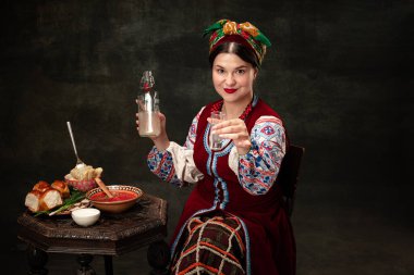 Greeting toast. Creative portrait of beautiful Ukrainian woman wearing traditional folk costume isolated over dark vintage background. Fashion, beauty, cultural heritage concept. Human emotions