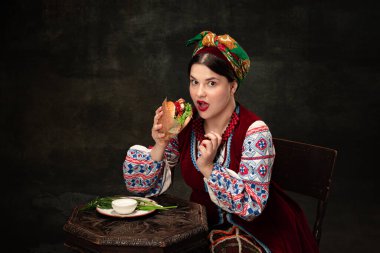 Tasting hamburger. One young beautiful Ukrainian woman in traditional folk Ukrainian costume isolated over dark vintage background. Fashion, beauty, cultural heritage, eras comparison concept.