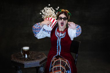 Screamer. Scared beautiful Ukrainian woman wearing traditional folk costume at movie theater isolated over dark vintage background. Fashion, beauty, cultural heritage concept. Human emotions