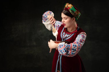 Making pancakes. Emotional young woman wearing national folk Ukrainian attire isolated over dark retro background. Human emotions, history, ad, fashion, beauty, eras comparison concept.