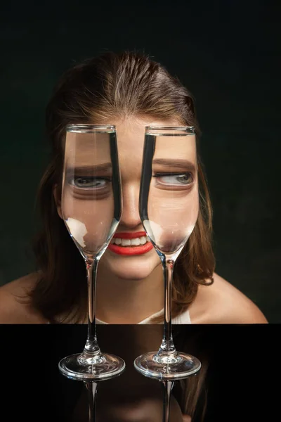 Joy, fun. Contemporary art with happy girl looking through wine glasses over dark background. Optical illusion. Concept of art, creativity, surrealism. Weird emotions, facial expression concept.