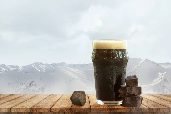 Dark chocolate and beer. Large glass of dark frothy beer standing on wooden table over snow-capped mountains background. Holidays, vacation, drinks, taste, ad and oktoberfest concept.
