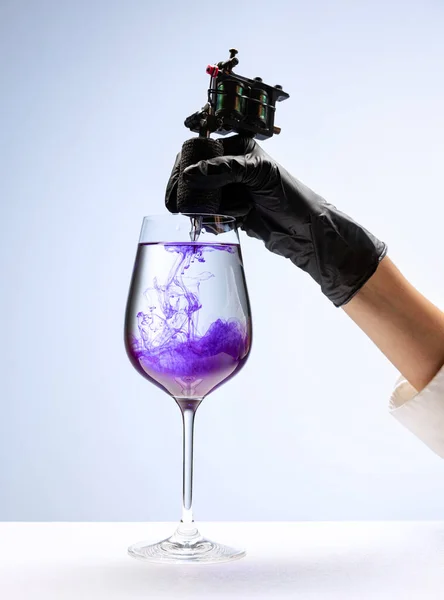 Wine glass and purple ink. Tattooer masters hand in black glove holding machine for making tattoo art on body isolated on white background. Concept of art, job, hobby, fashion, style
