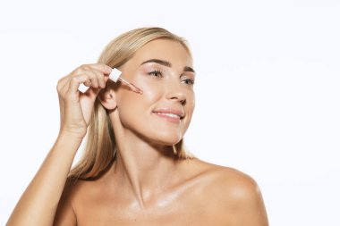 Gülümseyen genç bir kadın, beyaz arka planda yağ serumu uyguluyor. Kozmetik, makyaj, doğal ve ekolojik tedavi, cilt bakımı. Parlak ve sağlıklı cilt, moda, sağlık hizmetleri.