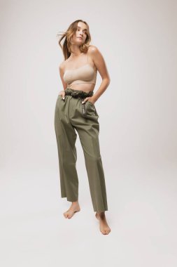 Uzun kıvırcık saçlı ve doğal makyajlı güzel bir kadın. Kırpık bluz giyiyor ve beyaz arka planda poz veren sıradan pantolonlar giyiyor. Kadın vücudunun doğal güzelliği, sanat, ilham, moda ve reklam.