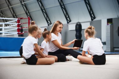 Koçluk. Acemi jimnastik sporcuları spor salonunda spor malzemeleriyle egzersiz yapıyorlar. Başarı kavramı, eğitim, hedefler. Bayan antrenör antrenörleri