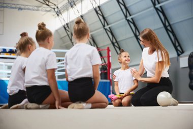 Çalışma ve eğitim metodu. Küçük kızlara öğretmenlik yapan genç bir kadın spor salonunda, iç mekanda yeni başlayan jimnastik sporcuları. Spor anlayışı, başarılar, öğrenim, hedefler, beceriler