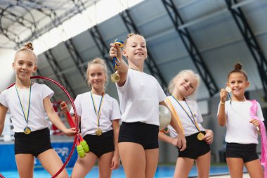 Mutlu küçük kızlar, bayan jimnastik sporcuları spor salonunda, kapalı alanda madalyalarını gösteriyorlar. Kazanma, spor, öğrenim, başarılar ve başarı kavramı. Takım çalışması, arkadaşlık ve destek