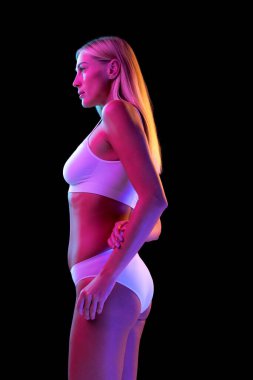 Profil görünümü. 35 yaşında güzel bir kadının portresi. Uzun sarı saçlı, beyaz iç çamaşırı giyen, neon ışıkta koyu renk arka planda izole edilmiş. Sağlıklı yaşam tarzı, cilt bakımı, güzellik, moda