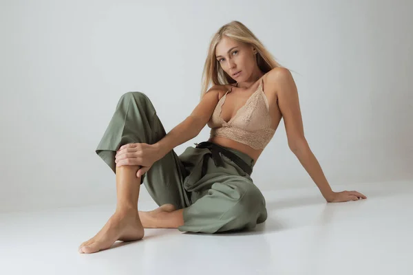 Beautiful woman with long blond hair and natural makeup wearing lacy top and casual trousers sitting on floor isolated on grey background. Natural beauty, art, inspiration, fashion and ad