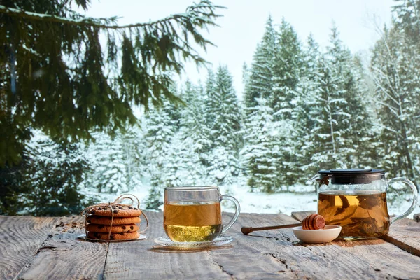 Winter cozy walks. Glass pot with herbal tea, cakes, biscuits on wooden table over winter snow forest background. Design for greeting card. Holidays, vacation, traditions