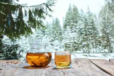 Kış yürüyüşleri. Kışın kar ormanı arka planında, ahşap masanın üzerinde bitki çayı bulunan cam çaydanlık. Tebrik kartı için tasarım, cihaz ekranı için duvar kağıdı. Tatiller, tatiller, gelenekler