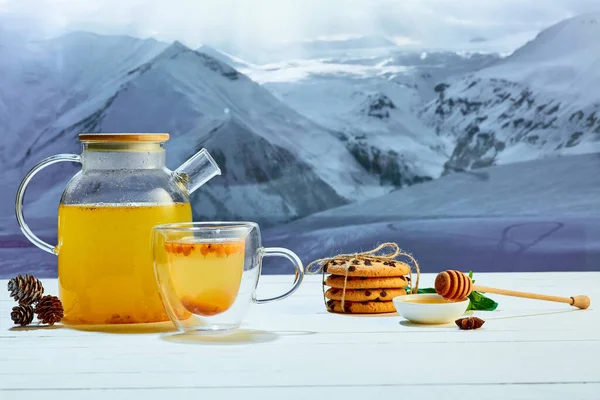 Seabuckthorn tea. Glass pot with fruit or berries tea, cakes, biscuits on table over winter snow natural landscape background. Design for greeting card, poster for ad. Holidays, vacation, traditions
