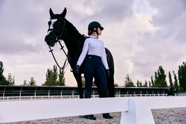 若いスポーツの女の子 プロのジョッキーや乗馬アリーナで黒馬とスポーツの制服やヘルメットで馬の女性 馬の衣装 乗馬だ スポーツ 優雅さ 広告の概念 — ストック写真