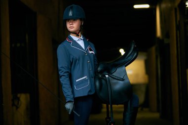 Genç kız, jokey ve at binicisi üniformalı ve kasklı kadın portresi at yarışı başlamadan önce ahırda duruyor. Binicilik sporu. At biniciliği. Spor, moda, reklam
