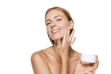 Yüzüne krem süren ve beyaz arka planda izole edilmiş kameraya bakan mutlu, yarı çıplak bir kızın güzel portresi. Spa, kozmetoloji, yaşlanma karşıtı tedavi kavramı