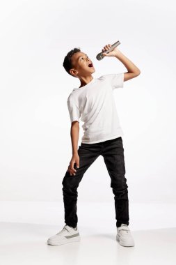 Müzik dersi. Beyaz arka planda izole edilmiş mikrofonla şarkı söyleyen küçük bir çocuğun portresi. İnsan duyguları, yüz ifadesi, sanat, müzik konsepti. İlan için alanı kopyala