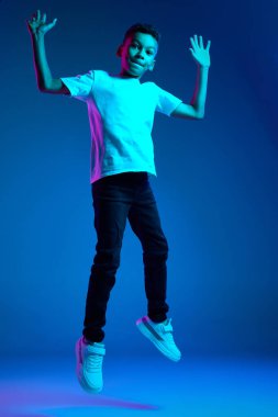Zıplıyorum, eğleniyorum. Duygusal küçük çocuğun stüdyo çekimi, farklı duyguları ifade eden çocuk mavi arka planda neon ışıkta izole edilmiş. Eğitim, çocukluk, modern yaşam tarzı