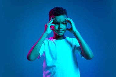 Çocuğun başı ağrıyor. Duygusal küçük çocuğun stüdyo çekimi, farklı duyguları ifade eden çocuk mavi arka planda neon ışıkta izole edilmiş. Eğitim, çocukluk, modern yaşam tarzı