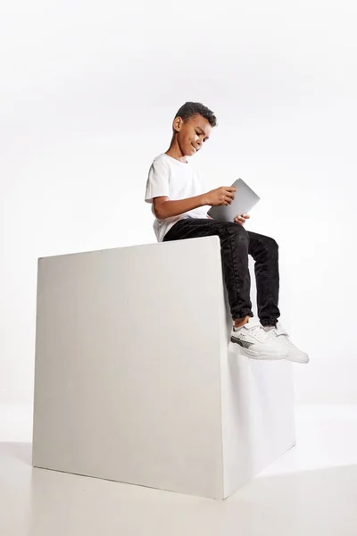 遠隔研究 白い背景にデジタルタブレットで大きな箱の上に座ってカジュアルなティーとジーンズで現在の学齢の少年 子供時代 子供のファッション — ストック写真