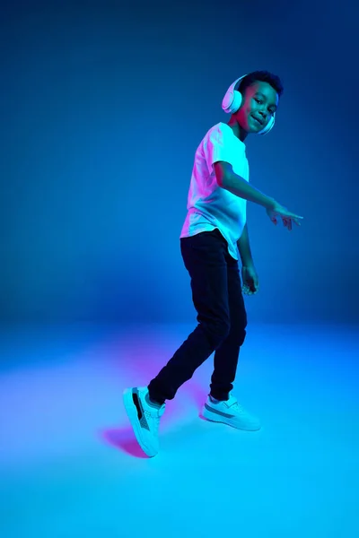 Dance. Little cheerful happy boy 6-7 years old wearing white t-shirt and headphones listen to music isolated on blue background in neon light. Concept of childhood, music, dance. Looks joyfull