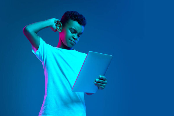 Remote education. Serious little boy, kid in white t-shirt looking at tablet screen isolated over blue background in neon light. Studying, gadgets, childhood, modern lifestyle